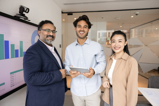 Three young and mature multiethnic professionals, analysts or business consultants look at camera standing in office next to digital screen displaying graphs and data presentation. Team collaboration