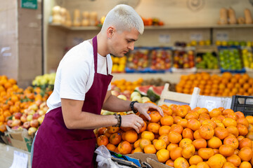 Young guy seller in apron puts fresh tangerines on display at vegetable market