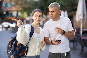 On pedestrian street, guy and girl are looking at smartphone screen, ordering taxi. Tourists use international taxi aggregator to call car..