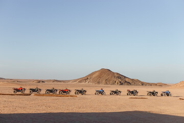 Group of men riders on ATV quad bike motorbikes driving through sandy barren desert landscape on sunny day scenic trip adventure tourism activity.