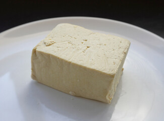 A piece of fresh tofu on a plate.
A whole block of fresh tofu sits on a white plate.  It is ideal for use in recipes of plant-based cuisine, dietary and vegan nutrition.