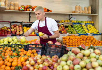 Young guy seller in apron puts fresh apples on display at vegetable market