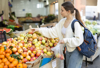 Woman with a backpack stands in the market and chooses fruits and ripe apples. Buying delicious fruits for a healthy lifestyle. Woman puts apples in plastic bag before weighing