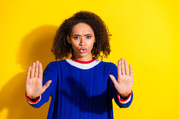 Confident young woman in blue sweater gesturing stop with hands against vibrant yellow background...