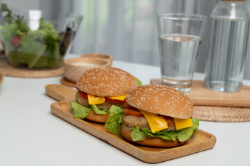 Focusing on double giant fried meat hamburger meal on square handle wooden board. Decorated by salad, homemade bun bread, beside drinking water on fast healthy food concept on white table. Infobahn.
