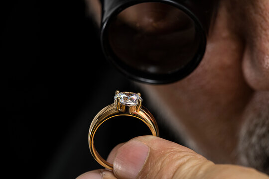 Jewelry appraiser examines a gold ring evaluating large diamond looking through magnifying glass monocular. Professional goldsmith manufacturing or repair luxury wedding engagement ring