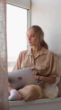 Young woman gets distance education via modern laptop sitting near window behind brown curtain in comfortable room at home slow motion