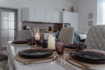 large dining table and upholstered grey chairs with backs. table is covered with linen tablecloth. dishes, glasses, candles on table. Dining room in a wooden country house. The interior of the house.