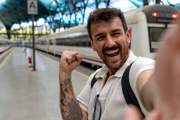 Excited man with backpack and tattoos taking a selfie at the train station platform after arriving...