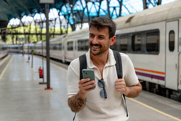 Happy tourist using mobile phone at railway station while waiting for train, holding his backpack