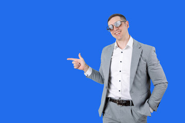 Successful young businessman in formal suit and sunglasses pointing finger at something on blue background. Young man pointing at something