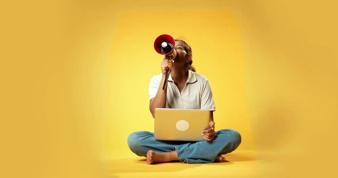 Girl Using Laptop with Megaphone While Sitting Against Yellow Seamless Background Making Online Announcement or Promotion, Smart Indian Asian Child Promoting Educational or Digital Content Loudly