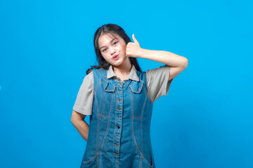 Young Asian woman in a denim dress showing a thumbs up gesture with a playful expression, standing in front of a solid blue background, symbolizing approval and positive vibes.