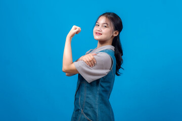 Fototapeta premium Confident young Asian woman smiling and flexing her bicep in a power pose against blue background. Her expression radiates empowerment, motivation, strength, and self-assurance.