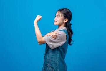 Fototapeta premium Confident young Asian woman smiling and flexing her bicep in a power pose against blue background. Her expression radiates empowerment, motivation, strength, and self-assurance.