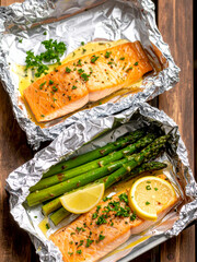 Baked salmon fillets with fresh herbs, lemon slices, and asparagus in aluminum foil on a rustic wooden table, healthy seafood dinner.