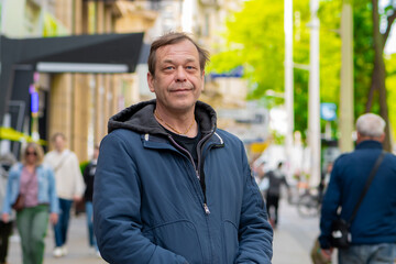 Obraz premium Portrait of an adult man aged 50-55 in a blue jacket standing on a busy city street, against a blurred background of passers-by and buildings.