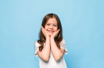 Fototapeta premium happy little brunette girl holding hands near face and smiling on blue background