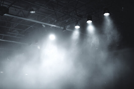 Dimly lit stage with spotlights casting beams of light through smoky atmosphere, creating dramatic and mysterious ambiance. industrial ceiling structure is visible above