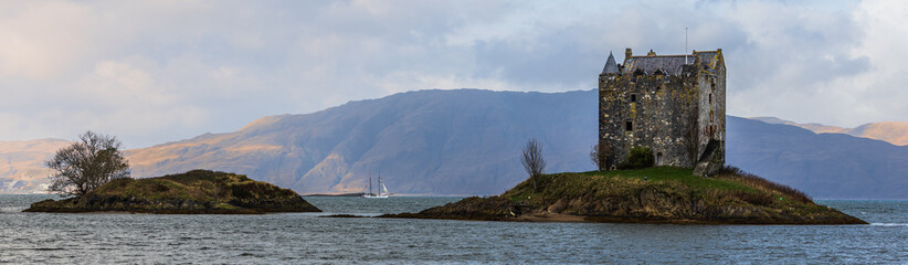 Castle Stalker an der schottischen Westk&uuml;ste mit Segelboot zwischen zwei kleinen Inseln, aufgenommen bei klarem Wetter in Argyll vor der ruhigen K&uuml;stenlandschaft von Loch Laich