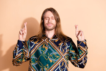 Young man with long brown hair crossing fingers and smiling in a patterned shirt against a beige...