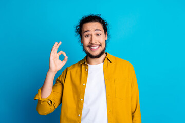 Cheerful young man in yellow shirt showing okay gesture against a solid blue background, embodying...