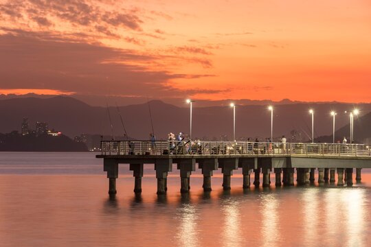 Santos city, Brazil. Colorful sunset on the fisherman's deck. Several people fishing and enjoying the day. In the background, the silhouette and lights of Porchat Island in the city of S&atilde;o Vicente.