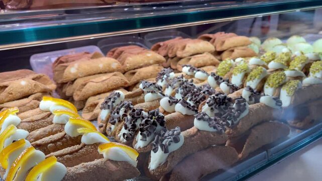 Assorted traditional Sicilian cannoli pastries filled with ricotta cream and decorated with chocolate chips, pistachios, and candied orange, displayed in a bakery glass case