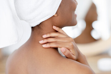 Beauty and spa day at home at weekends and free time. Pretty fresh millennial african american woman in white towel smears her body with cream after shower, in interior of bedroom in morning, back