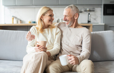 A happy senior couple sits closely on a sofa, sharing coffee and smiles in a stylish kitchen...