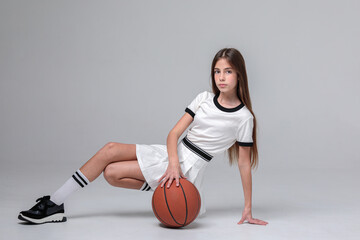 Naklejka premium Beautiful teenage girl with long hair in a white sports skirt and top with a basketball in a photo studio on a white background. Sportswoman. Basketball. Youth.