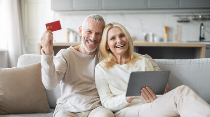 A joyful elderly couple relaxes on their couch, smiling while holding a credit card and tablet, engaged in online shopping during a cozy evening at home.