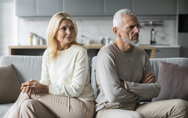 A senior couple appears emotionally distant as they sit on a couch in their living room,...