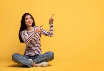 Asian young woman with long hair sits cross-legged on a bright yellow background, happily pointing...