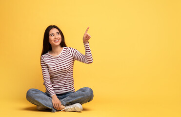 Asian young woman with long hair sits cross-legged on a bright yellow background, smiling and...