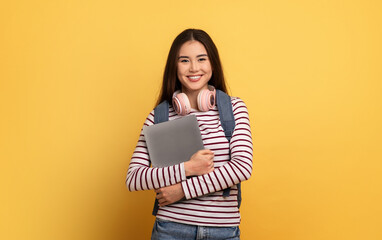 A cheerful young woman stands against a bright yellow background, holding a laptop close to her chest. She wears headphones around her neck and a striped top, exuding confidence and warmth.