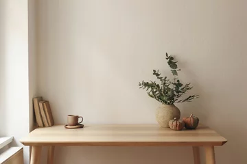 Fotobehang Scandinavisch Autumn interior still life. Vase with eucalyptus, olive tree branches, pumpkins. Cup of tea, coffee, books. Wooden table at window. Home office. Minimal Scandi interior. Empty white wall background.  © tabitazn