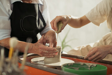 Potter using sponge helping senior woman creating clay vessel on pottery wheel