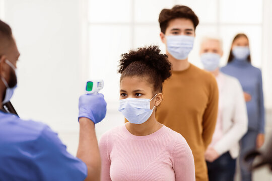 Temperature Screening Concept. Nurse Measuring Fever For Black Patient Girl Standing In Queue Line, Using Infrared Contactless Themometer In Hospital Waiting-Room. Coronavirus Covid-19 Protection