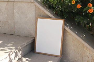 Fototapeta na wymiar Vertical wooden picture frame leaning against stone stairway in sunlight. Textured beige tiles background, orange flowers, green leaves. Summer outdoor natural, minimal backdrop. Empty poster mockup.