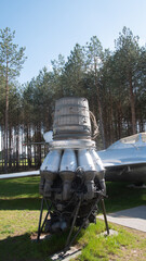 A detailed view of a vintage Soviet rocket engine showcased outdoors near an aircraft, surrounded by lush pine trees under a clear blue sky.