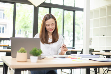 Obraz premium A woman is working at a desk in an office with a laptop, a mobile phone and a pile of documents. She is smiling and happy. 