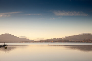 Fototapeta premium Golden dusk settles over Lake Worthersee near Klagenfurt; Austria. calm mirror-like water reflects distant wooded hills and pale Alpine silhouettes beneath a deepening blue sky.
