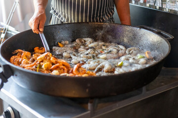 Seafood is being cooked in a large pan at a lively outdoor market during a warm afternoon
