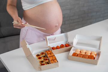 A pregnant woman holds chopsticks and stands at the table with rolls. Close-up of the belly.