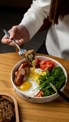 Dining at a charming cafe, a person enjoys a hearty breakfast bowl filled with sunny-side-up eggs, crispy sausages, fresh tomatoes, and vibrant greens, evoking a sense of comfort and indulgence