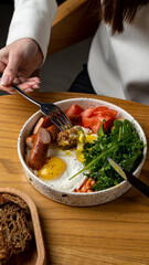 A delightful breakfast bowl is presented, showcasing perfectly cooked eggs, savory sausage, fresh tomatoes, and vibrant greens. The wooden table adds a rustic charm to the meal