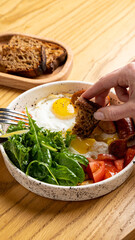 A hand dips a piece of toasted bread into a colorful bowl filled with eggs, sausage, fresh greens, and tomatoes, showcasing a vibrant breakfast masterpiece. Enjoying the rich flavors and textures