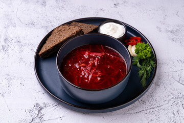 A bowl of rich red borscht brimming with vegetables sits on a dark plate, accompanied by slices of rye bread and a dollop of sour cream, creating a warm, inviting atmosphere