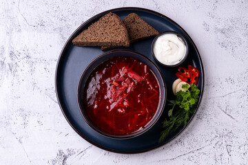 A vibrant bowl of borscht, garnished with fresh herbs, sliced red chili, and a dollop of sour cream, sits next to hearty rye bread on a textured surface. An inviting meal awaits